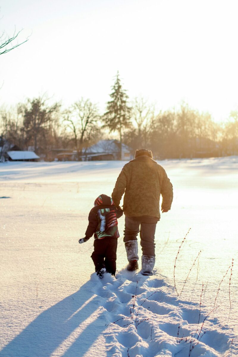 5 fun and festive winter family photo ideas to try this season with man holding a childs hand walking in the snow with footprints follow behind